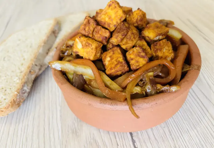 Tofu in padella dorato e saporito, servito con cipolle caramellate in una ciotola di terracotta, accanto a fette di pane rustico.