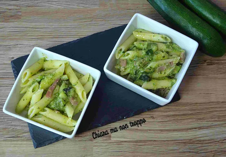 Due ciotole di pasta penne con zucchine e pancetta, condite con un pesto verde. Due zucchine fresche riposano sullo sfondo.