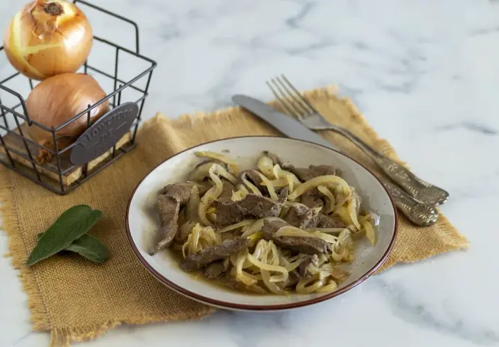 Piatto di fegato alla veneziana con cipolle dorate, accompagnato da cipolle fresche in un cestino e foglie di salvia.