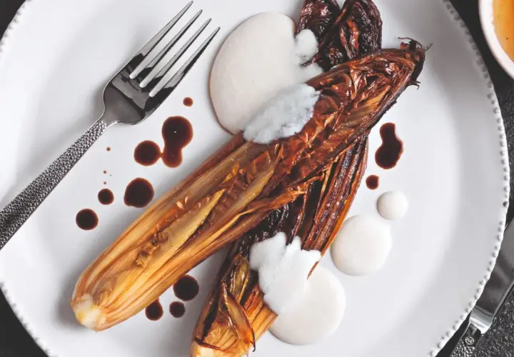Radicchio al forno caramellato, servito con crema bianca e salsa scura su un piatto bianco, con forchetta e coltello.