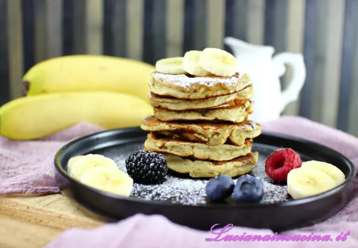 Pancakes alla banana soffici e dorati, guarniti con fettine di banana, frutti di bosco e zucchero a velo.