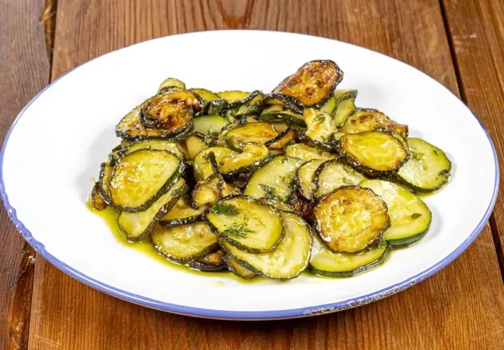 Zucchine alla poverella, fette sottili e dorate, condite con olio e erbe aromatiche, su un piatto bianco con bordo blu.