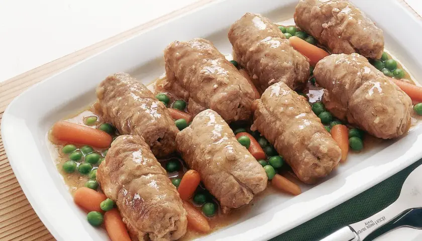 Sette involtini di carne succulenti in un piatto bianco con piselli e carote.
