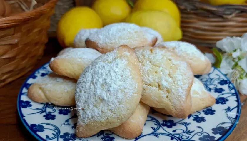 Biscotti con farina di mandorle spolverati di zucchero a velo, su un piatto decorato con fiori blu. Sullo sfondo limoni e un cestino.