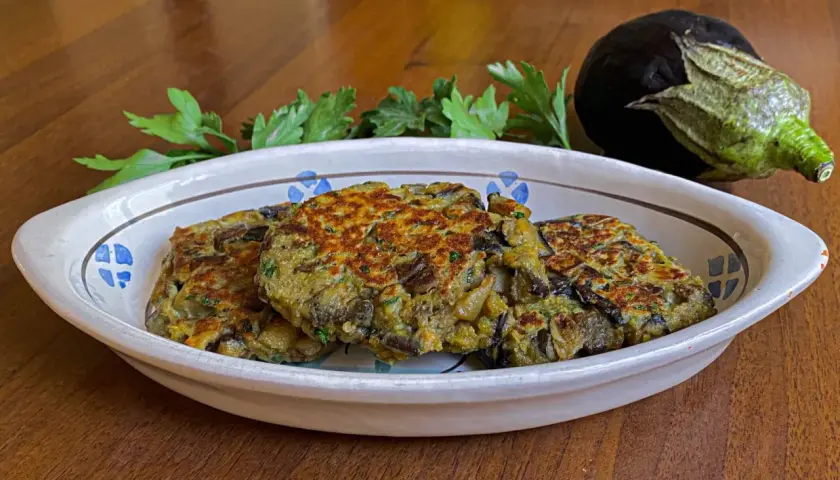 Tre hamburger di melanzane dorati e appetitosi in un piatto decorato, con un'altra melanzana e prezzemolo sullo sfondo.