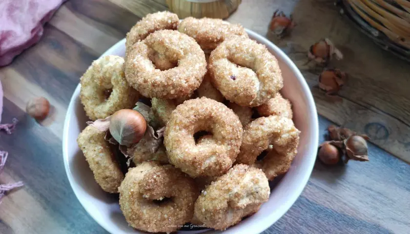 Ciambelline al vino cosparse di zucchero, con nocciole e un drappo rosa su un tavolo di legno.