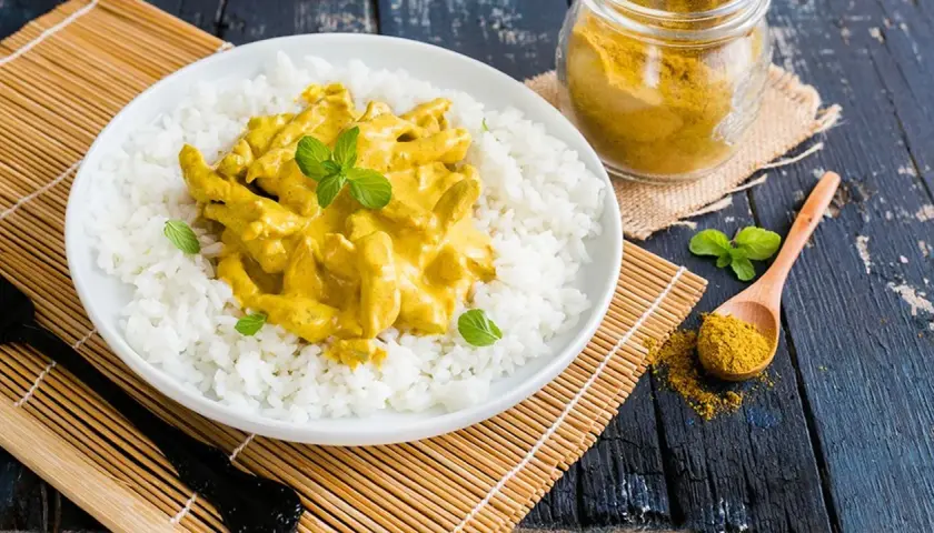 Piatto di riso bianco con straccetti di pollo al curry cremoso, guarnito con menta fresca. Un cucchiaio di curry in polvere e un vasetto completano la scena.