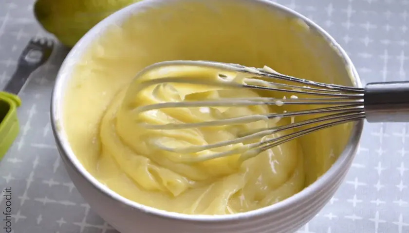 Una ciotola bianca piena di soffice crema al limone, con una frusta che la sta mescolando. Sullo sfondo, un limone intero.