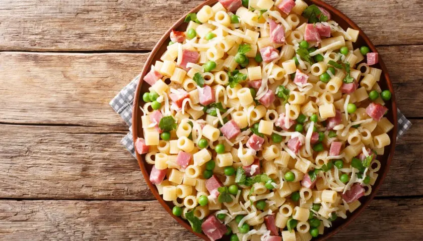Un piatto di pasta con prosciutto cotto, piselli e formaggio grattugiato su un tavolo di legno rustico.