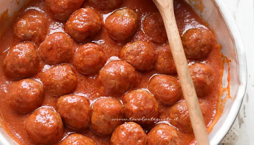 Polpette al sugo succulente in una ciotola bianca, pronte per essere gustate. Un cucchiaio di legno le accompagna.