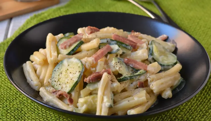 Piatto di pasta cremosa con zucchine a rondelle e cubetti di speck croccante, guarnito con pepe nero macinato.