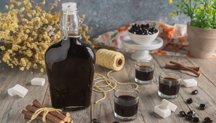 Bottiglia di liquore alla liquirizia scuro, bicchierini, bastoncini di liquirizia, zollette di zucchero e caramelle nere su un tavolo di legno.