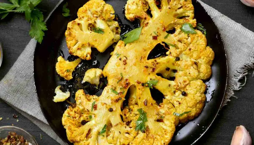 Fette di cavolo al forno dorate e speziate, servite su una padella nera con erbe fresche e pepe nero.