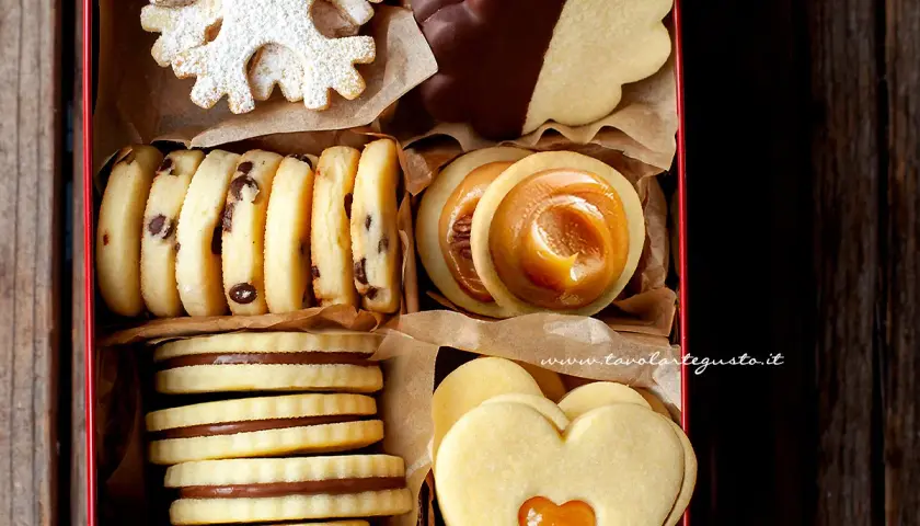Scatola piena di biscotti classici: a fiocco di neve, con gocce di cioccolato, ripieni di caramello, a forma di cuore e ricoperti di cioccolato.
