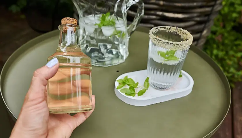 Mano tiene una bottiglietta di sciroppo di menta, accanto a un bicchiere e una caraffa con acqua, ghiaccio e foglie di menta.