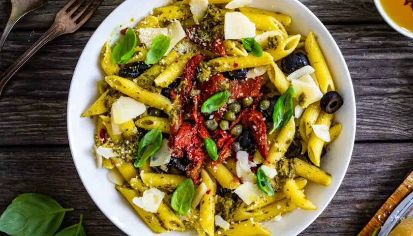 Penne al pesto con pomodori secchi, olive nere, capperi e scaglie di parmigiano. Un piatto colorato e saporito.