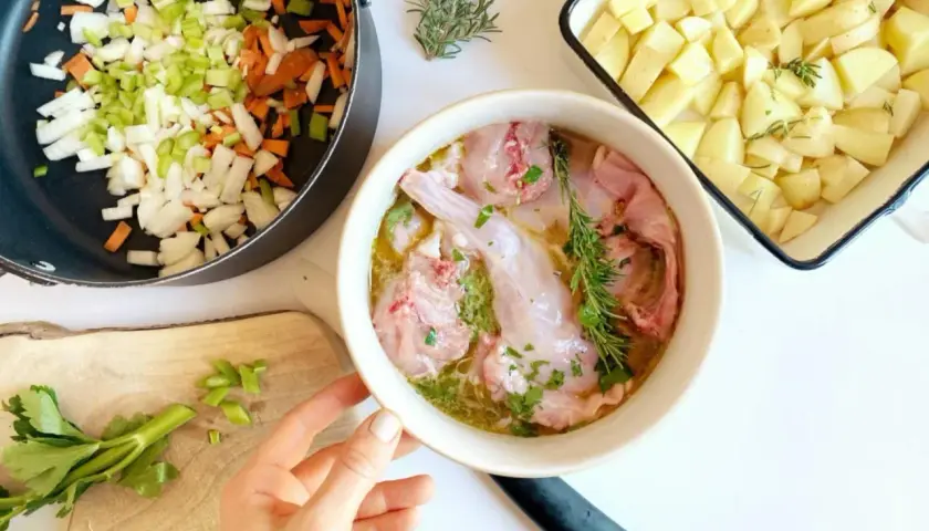 Preparazione per un coniglio alla cacciatora in bianco: pezzi di coniglio crudo in una ciotola, verdure tritate in padella e patate a cubetti in una teglia.