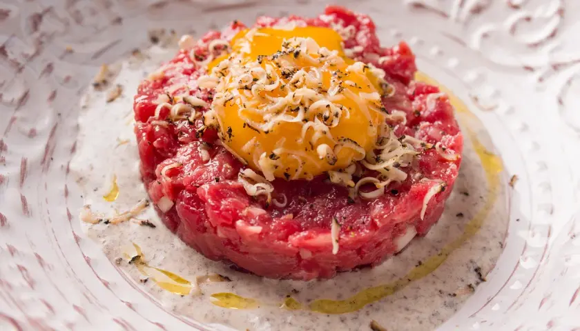 Deliziosa tartare di manzo finemente tritata, guarnita con tuorli d'uovo dorati e scaglie di formaggio, servita su un piatto elegante con una salsa cremosa.