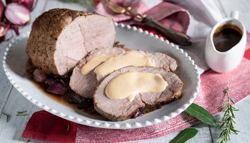 Arrosto di vitello morbido con sughetto benedetta, servito su un piatto bianco con cipolle rosse e rosmarino.