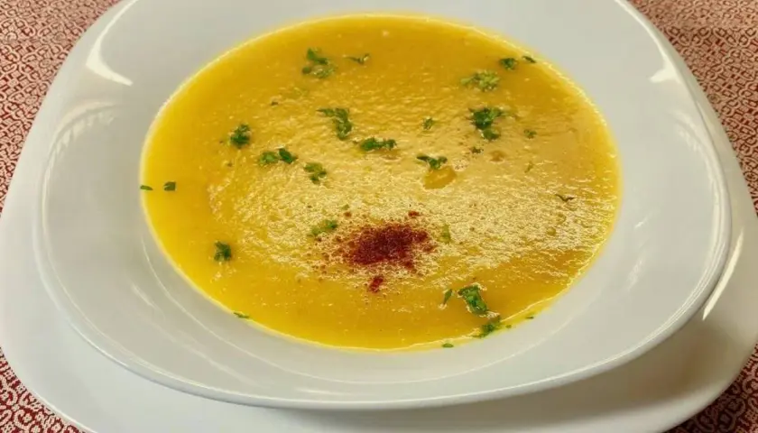 Una vellutata crema di peperoni, guarnita con prezzemolo fresco e una spolverata di paprika, servita in una ciotola bianca su un tovagliolo rosso.