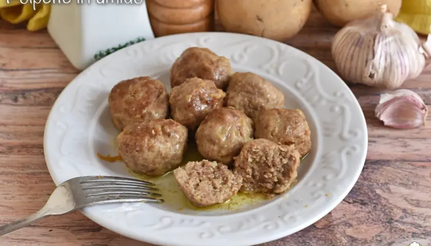 Polpette in umido succulente su un piatto bianco, con una forchetta pronta ad assaggiarle. Sullo sfondo, patate e aglio.