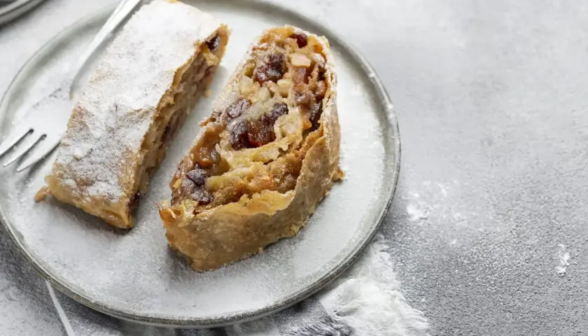 Due fette di strudel di mele con pasta sfoglia, cosparse di zucchero a velo, su un piatto grigio con una forchetta.