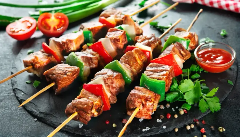 Spiedini di carne succulenti alternati a peperoni rossi e verdi, cipolla bianca, serviti su una pietra scura con pomodorini e salsa.