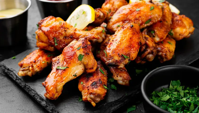 Alette di pollo dorate e succulente, guarnite con prezzemolo fresco e spicchi di limone, servite su una pietra scura con salse a lato.