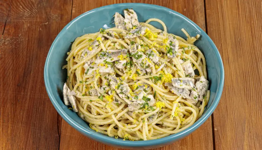 Spaghetti con acciughe, prezzemolo tritato e scorza di limone grattugiata in una ciotola blu su un tavolo di legno.