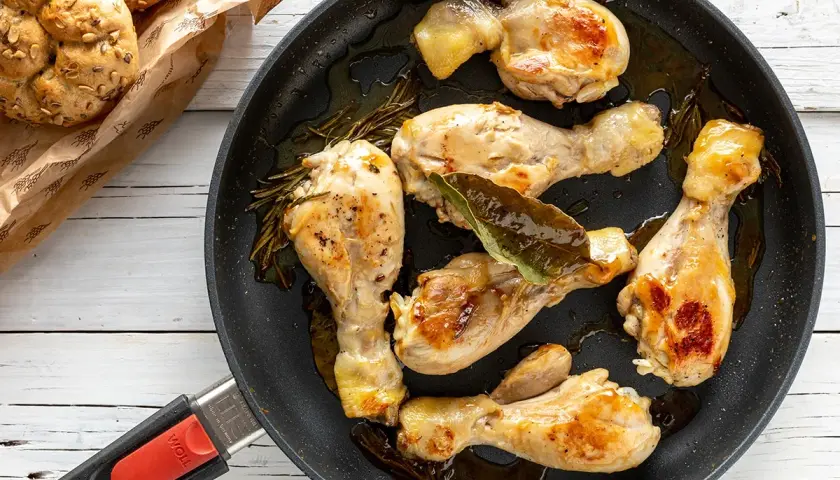 Fusi di pollo in padella, dorati e succulenti, con rosmarino e alloro. Accanto, panini integrali appena sfornati.