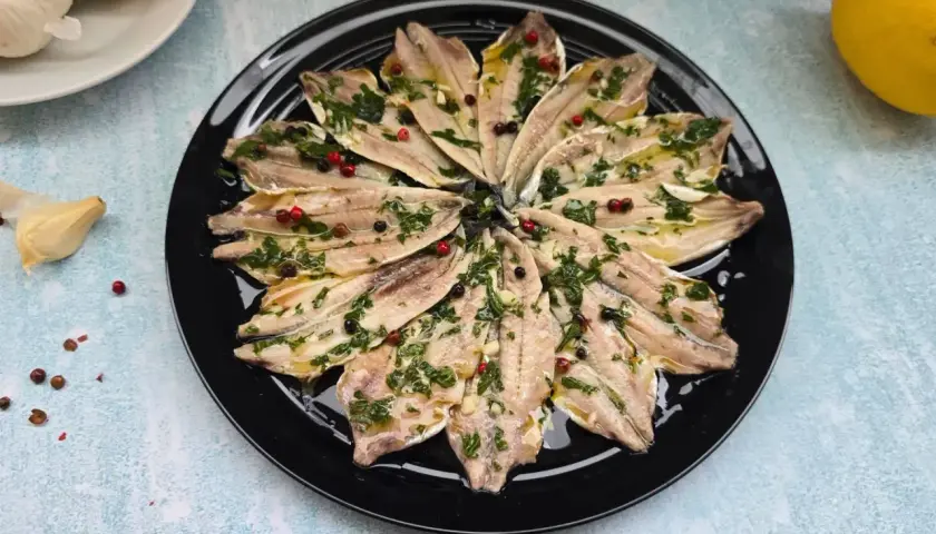 Alici marinate ricetta: filetti di acciughe conditi con erbe aromatiche, aglio e pepe in grani su un piatto nero.