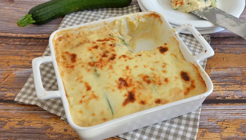 Parmigiana di zucchine bianca senza besciamella, dorata e filante, pronta per essere gustata. Accanto, una zucchina fresca.