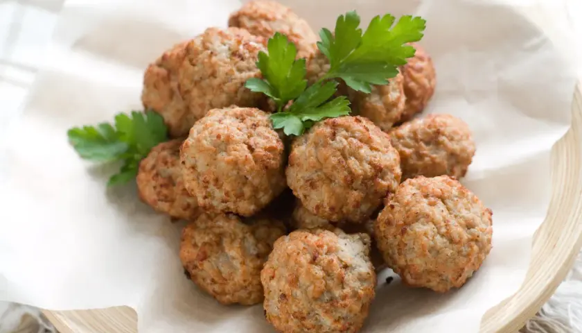 Un mucchio di polpette di pollo dorate e profumate, guarnite con prezzemolo fresco, servite su carta da forno in un piatto di legno.