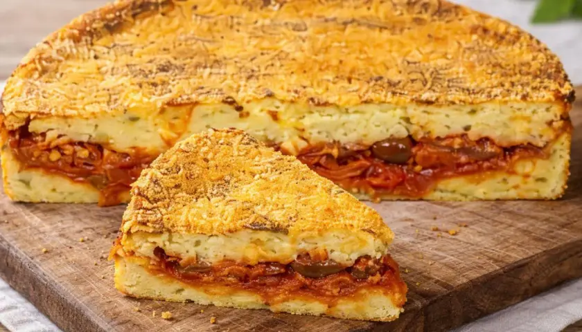 Una fetta di pitta di patate, con strati di purè e un ricco ripieno di pomodoro e olive, su un tagliere di legno.