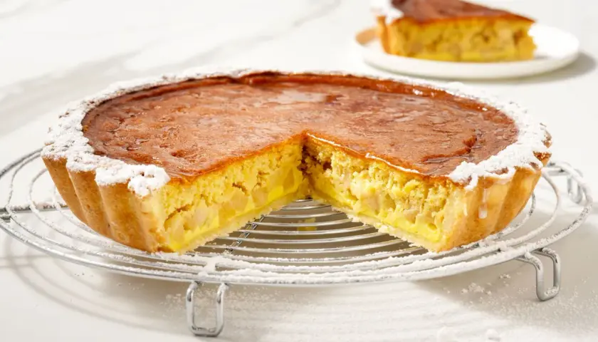 Torta di mele con crema pasticcera, con una fetta tagliata, cosparsa di zucchero a velo su una griglia metallica.
