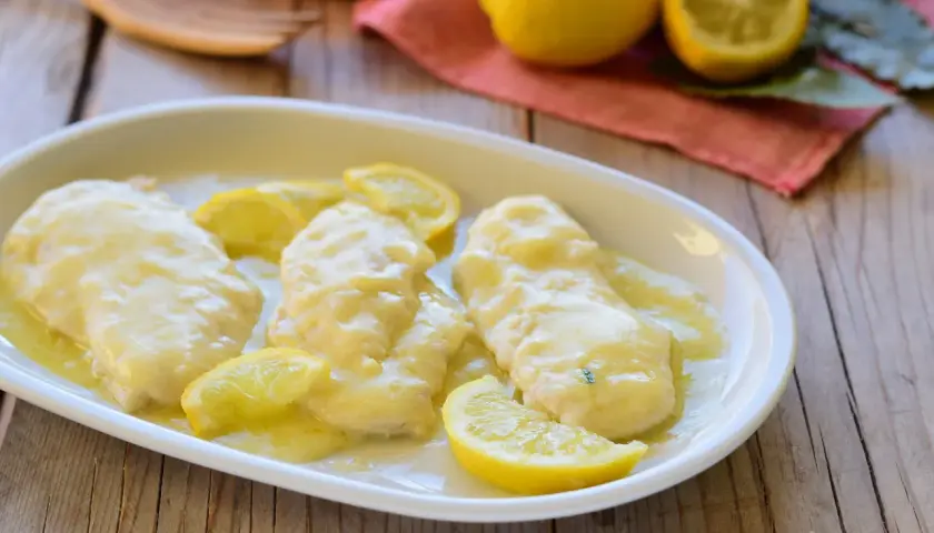 Scaloppine al limone cremose, succulente e profumate, guarnite con spicchi di limone fresco su un piatto bianco.