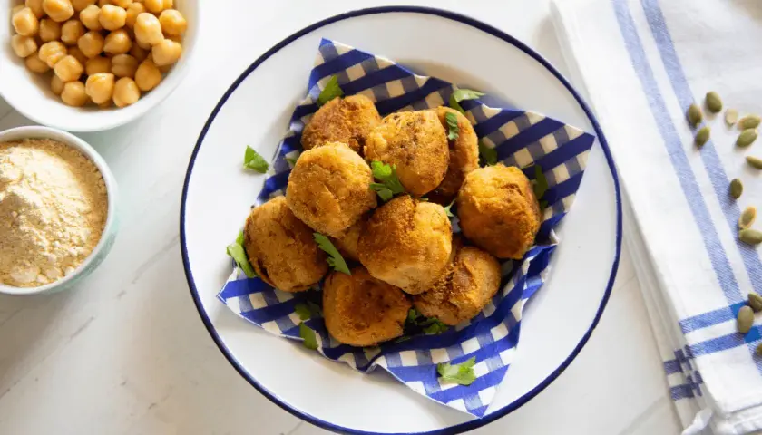 Piatto di polpette di ceci dorate e croccanti, guarnite con prezzemolo fresco. Accanto, ciotole con ceci e farina di ceci.