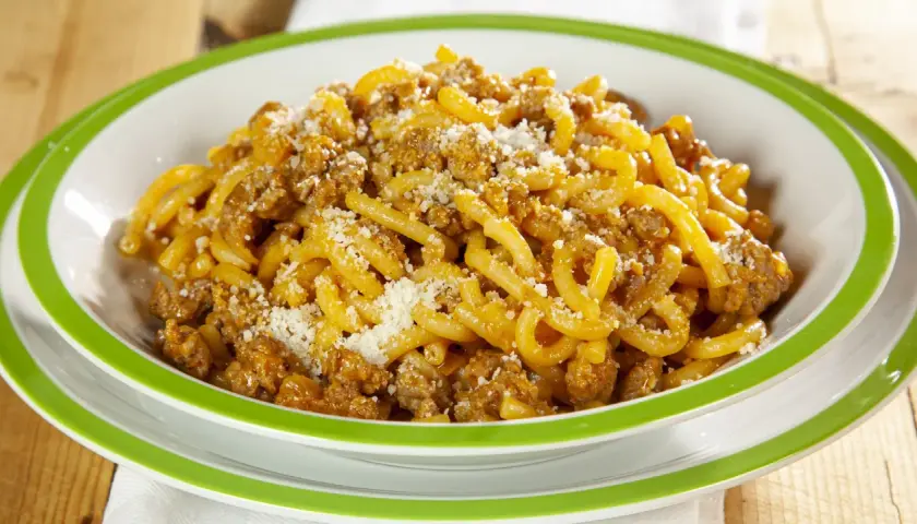 Piatto di gramigna alla salsiccia, condita con ragù e parmigiano grattugiato, servita in una ciotola bianca con bordo verde.