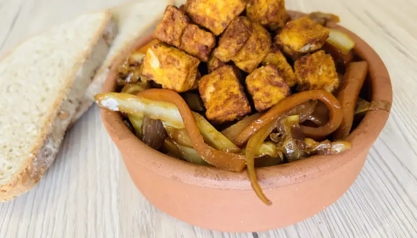 Tofu in padella dorato e saporito, servito con cipolle caramellate in una ciotola di terracotta, accanto a fette di pane rustico.