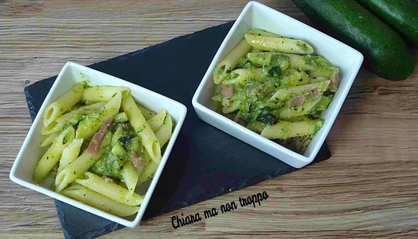 Due ciotole di pasta penne con zucchine e pancetta, condite con un pesto verde. Due zucchine fresche riposano sullo sfondo.