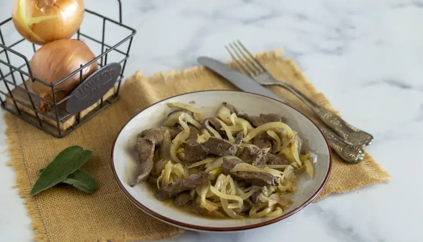 Piatto di fegato alla veneziana con cipolle dorate, accompagnato da cipolle fresche in un cestino e foglie di salvia.