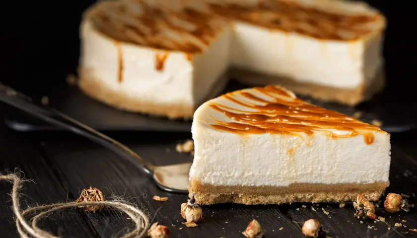Una fetta di cheesecake senza gelatina, con caramello colante, su un tavolo di legno scuro.