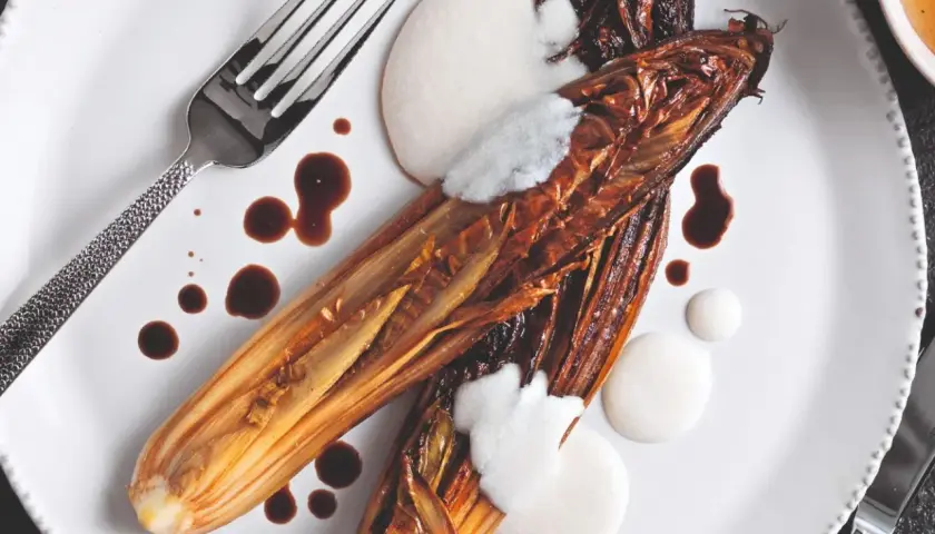 Radicchio al forno caramellato, servito con crema bianca e salsa scura su un piatto bianco, con forchetta e coltello.