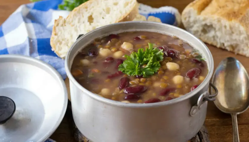 Una fumante zuppa di legumi, ricca di fagioli rossi, lenticchie e ceci, guarnita con prezzemolo fresco, accanto a pane casereccio.