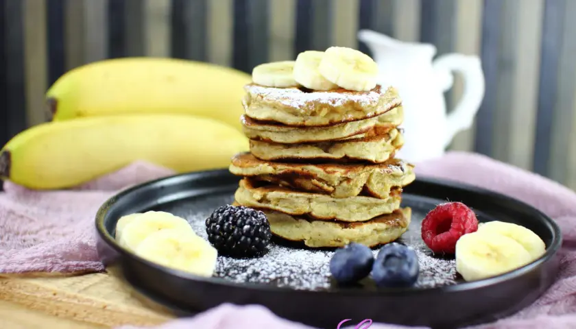 Pancakes alla banana soffici e dorati, guarniti con fettine di banana, frutti di bosco e zucchero a velo.