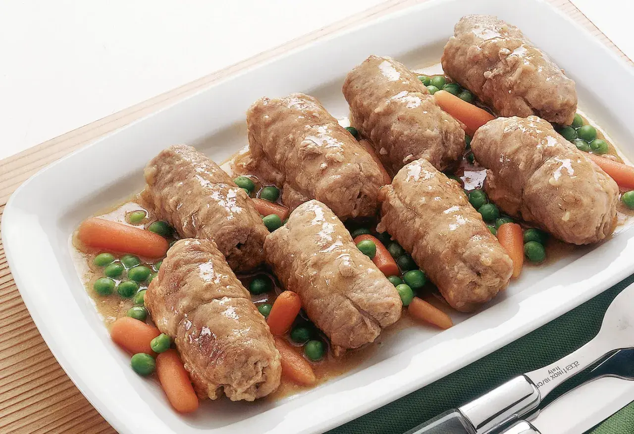 Sette involtini di carne succulenti in un piatto bianco con piselli e carote.