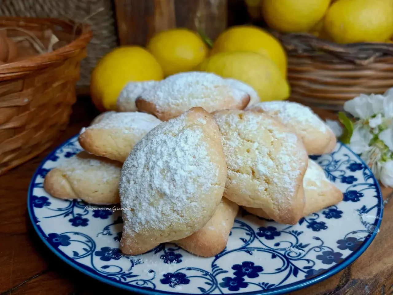 Biscotti con farina di mandorle spolverati di zucchero a velo, su un piatto decorato con fiori blu. Sullo sfondo limoni e un cestino.