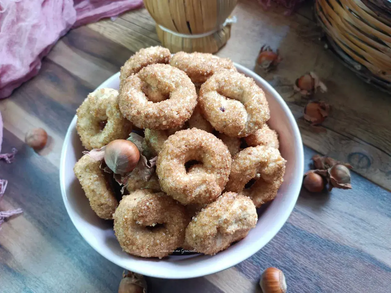 Ciambelline al vino cosparse di zucchero, con nocciole e un drappo rosa su un tavolo di legno.