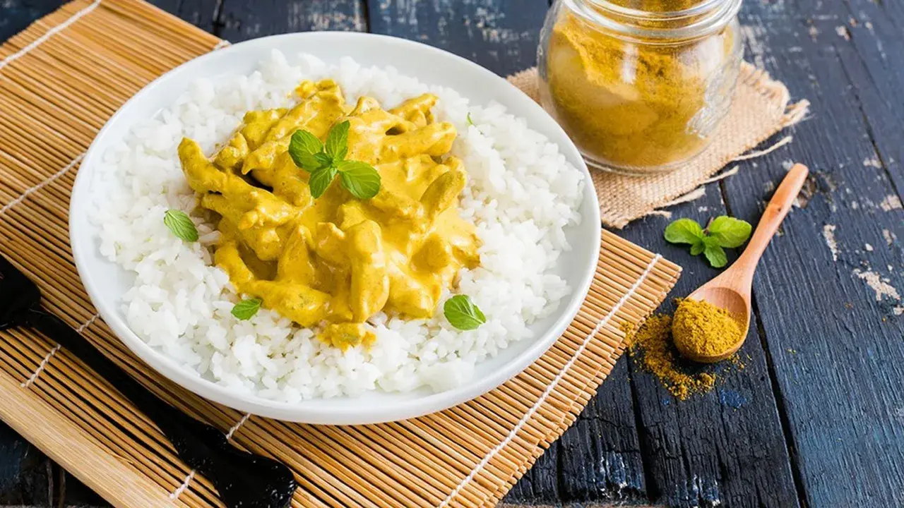 Piatto di riso bianco con straccetti di pollo al curry cremoso, guarnito con menta fresca. Un cucchiaio di curry in polvere e un vasetto completano la scena.