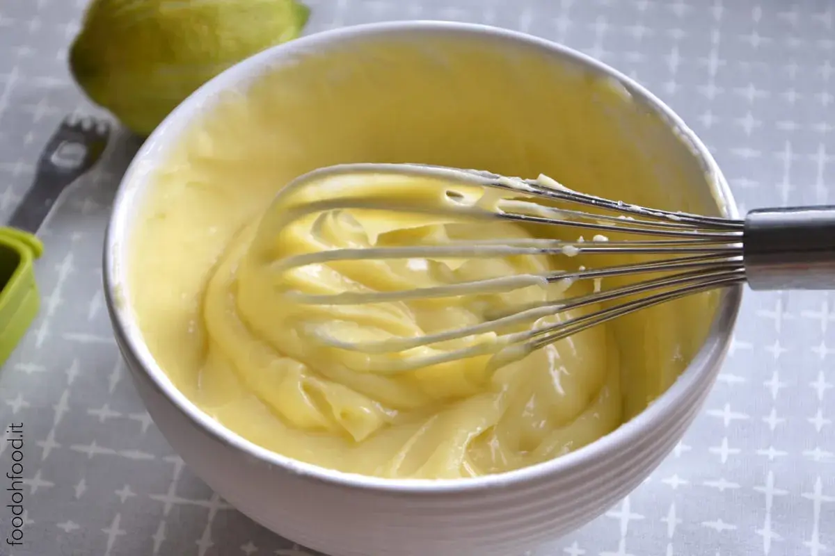 Una ciotola bianca piena di soffice crema al limone, con una frusta che la sta mescolando. Sullo sfondo, un limone intero.